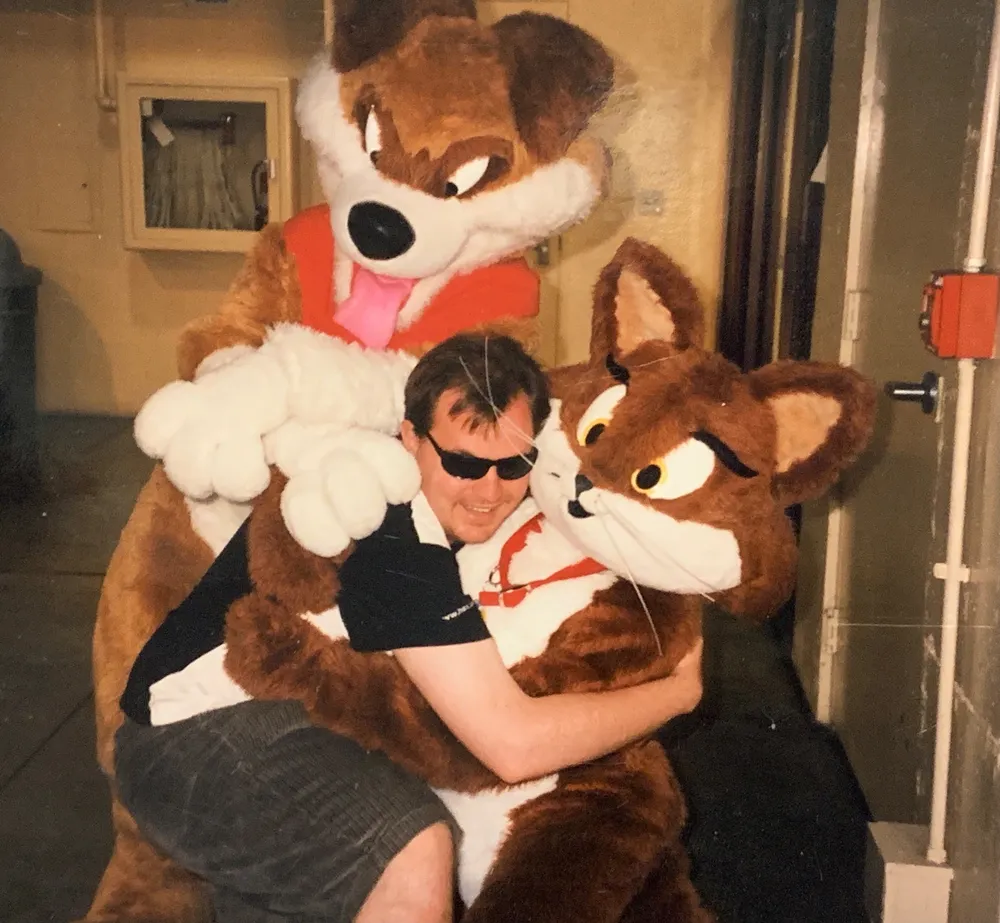 A picture of me, then a masc-presenting 25 year old, wearing sunglasses and board shorts, joyfully hugging two fursuiters, at a California furry convention